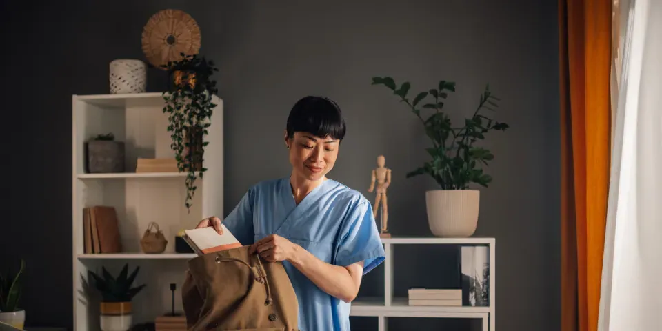 Asian dedicated nurse in blue scrubs is seen preparing medical tools and organizing documents into a brown backpack in a well-lit, modern room with shelves and assorted decor.